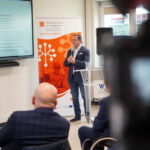 Prelegent z mikrofonem prowadzący prezentację przed uczestnikami spotkania w sali konferencyjnej