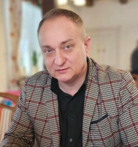 Portret mężczyzny w średnim wieku, Adama Skorniewskiego, ubranego w elegancką marynarkę w czerwoną kratę i czarną koszulę. Mężczyzna patrzy prosto w obiektyw, siedząc w pomieszczeniu z rozmytym tłem.