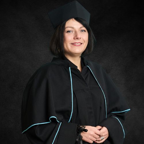 Portret uśmiechniętej kobiety w czarnej todze akademickiej z jasnoniebieskimi lamówkami i czarnym birecie. Kobieta ma ciemne włosy i patrzy prosto w obiektyw, a jej dłonie są złożone z przodu.