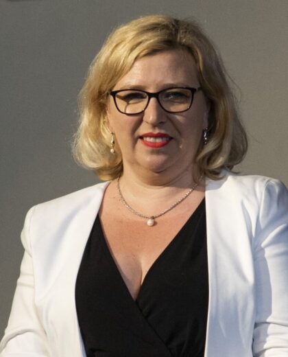 Portret kobiety w okularach, białej marynarce i czarnej bluzce na szarym tle