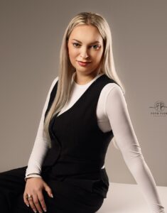 Młoda kobieta z długimi blond włosami, ubrana w białą bluzkę z długim rękawem i czarną kamizelkę, siedzi i patrzy w obiektyw z delikatnym uśmiechem.