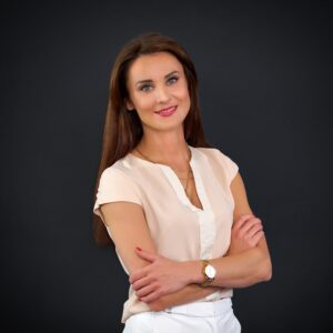 Portret uśmiechniętej kobiety o długich brązowych włosach, ubranej w jasną bluzkę i białe spodnie, stojącej z założonymi rękami na ciemnym tle. Kobieta ma na sobie zegarek i delikatny makijaż.