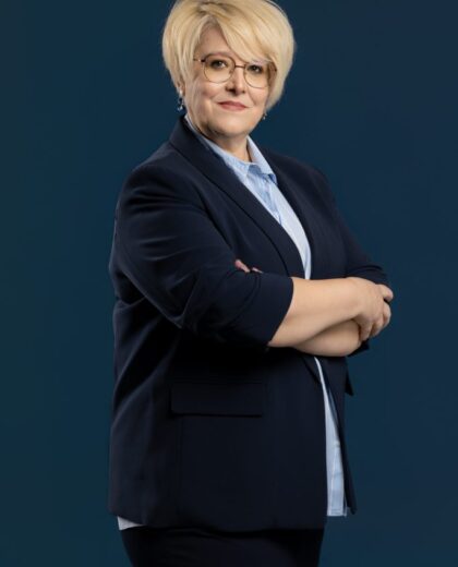 Kobieta z krótkimi blond włosami i okularami, ubrana w granatową marynarkę i jasnoniebieską koszulę, stoi ze skrzyżowanymi ramionami na ciemnoniebieskim tle.
