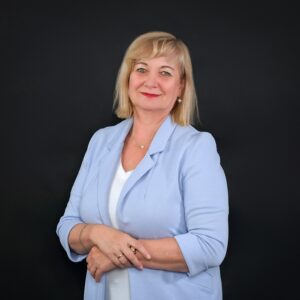 Portret uśmiechniętej kobiety z blond włosami, ubranej w jasnoniebieską marynarkę i białą bluzkę. Kobieta ma skrzyżowane ręce i patrzy wprost na widza, stojąc na jednolitym, ciemnym tle.