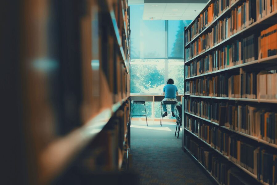 Osoba siedząca tyłem przy biurku z dużym oknem w tle, otoczona regałami pełnymi książek w bibliotece. Użytkownik ma na sobie słuchawki i skupia się na pracy lub nauce.
