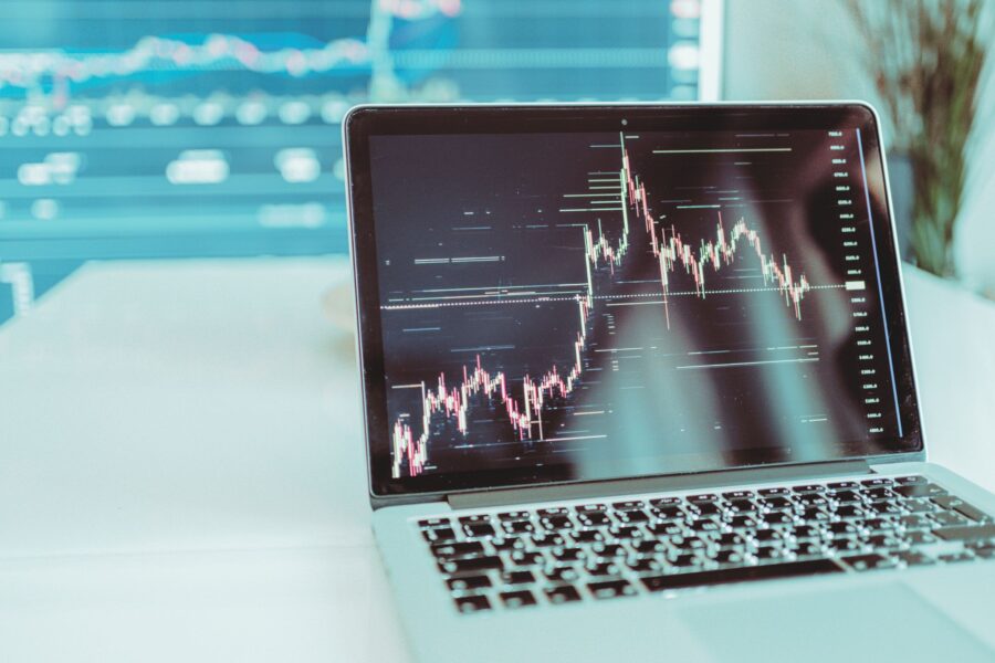 Laptop z otwartym ekranem, na którym wyświetlany jest wykres świecowy przedstawiający dane finansowe lub giełdowe. W tle widoczny jest rozmyty drugi monitor z podobnymi wykresami.