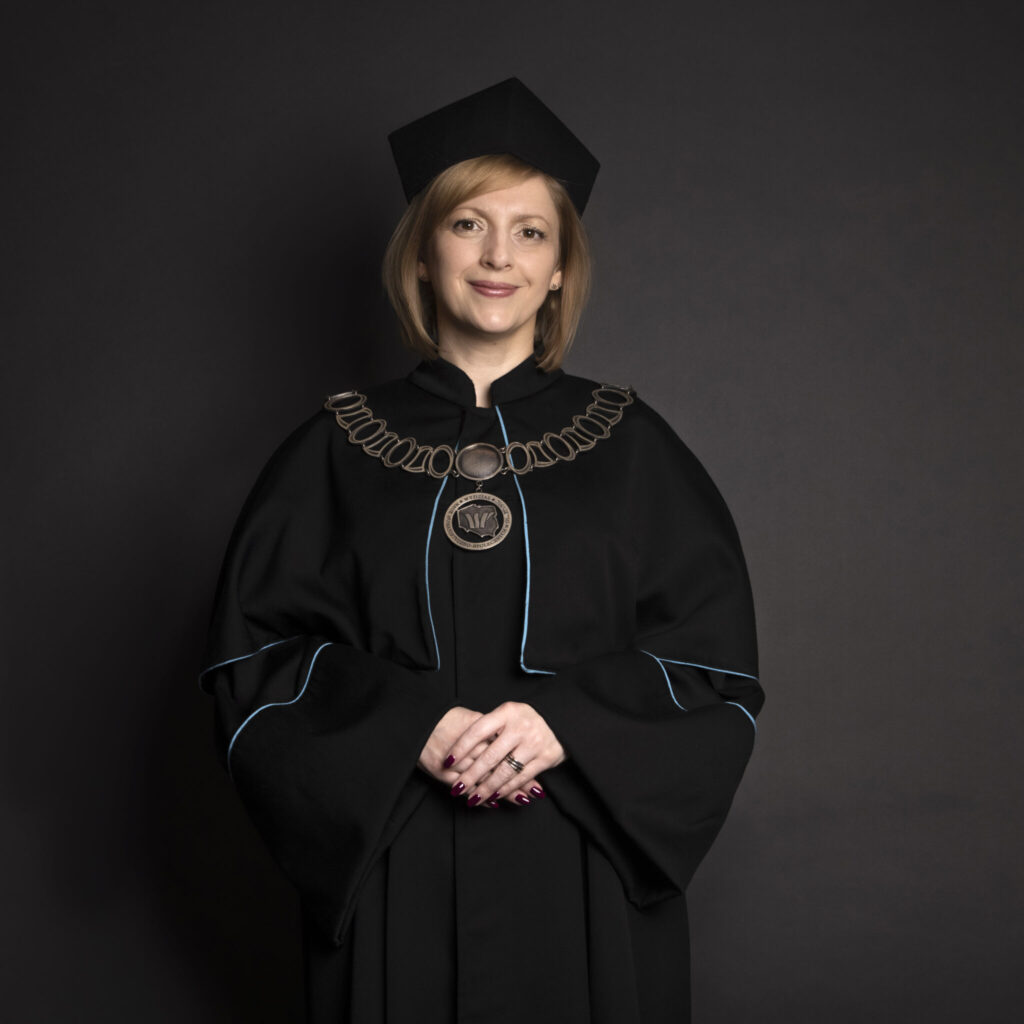 Portret uśmiechniętej kobiety w czarnej todze akademickiej z niebieskimi akcentami i birecie. Na szyi ma ceremonialny łańcuch z medalionem Wydziału Humanistyczno-Społecznych, a ręce ma złożone z przodu.