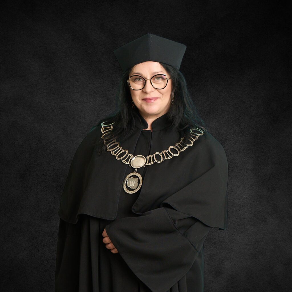Portret uśmiechniętej kobiety w okularach, ubranej w czarną togę akademicką i biret. Na szyi ma łańcuch dziekański z medalionem Wydziału Zamiejscowego w Wałbrzychu.