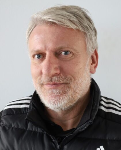 Portret mężczyzny z siwymi włosami i brodą w czarnej kurtce na jasnym tle
