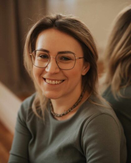 Portret uśmiechniętej kobiety w okularach i zielonej bluzce w jasnym pomieszczeniu