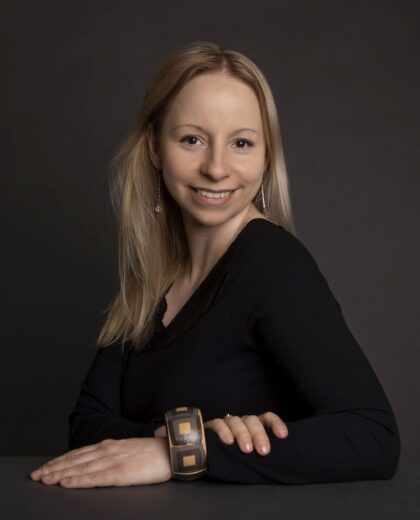 Młoda kobieta z blond włosami i uśmiechem, ubrana w czarną bluzkę, siedzi przy stole z rękami opartymi na blacie. Na jej lewym nadgarstku widoczna jest ozdobna bransoletka.