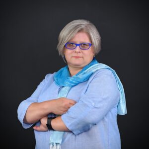 Portret Alicji Pazur, kobiety z siwymi włosami i niebieskimi okularami, ubranej w błękitną koszulę i szal, stojącej ze skrzyżowanymi ramionami na ciemnym tle.