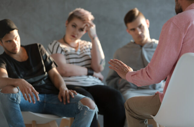 Grupa młodych ludzi, w tym dwóch mężczyzn i jedna kobieta, siedzi w kręgu podczas sesji terapeutycznej lub spotkania socjoterapeutycznego. Osoba prowadząca, widoczna od tyłu, gestykuluje ręką, a uczestnicy słuchają z uwagą.