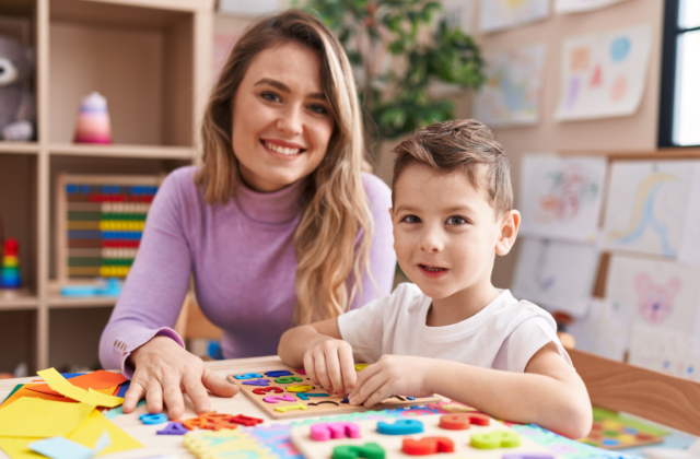 Uśmiechnięta kobieta i mały chłopiec siedzą przy stole, bawiąc się edukacyjnymi puzzlami z cyframi. Na stole leżą kolorowe materiały plastyczne, a w tle widać półki z zabawkami i rysunki dzieci, co sugeruje salę lekcyjną lub pokój zabaw.