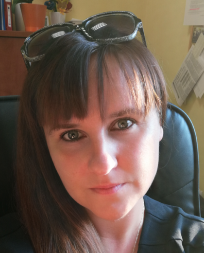 Portret kobiety z długimi brązowymi włosami i grzywką, siedzącej w biurze. Ma na głowie okulary przeciwsłoneczne i patrzy prosto w obiektyw.