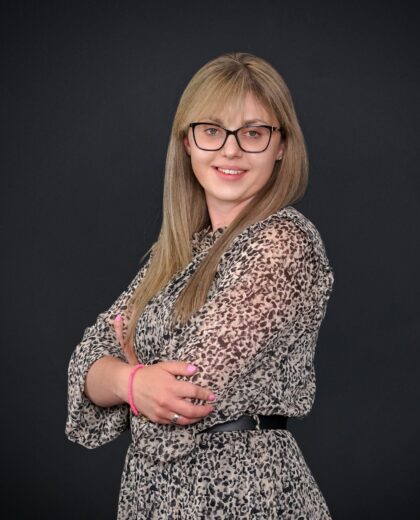 Młoda kobieta z długimi blond włosami i okularami, ubrana w sukienkę w panterkę, uśmiecha się, stojąc ze skrzyżowanymi ramionami na ciemnym tle.