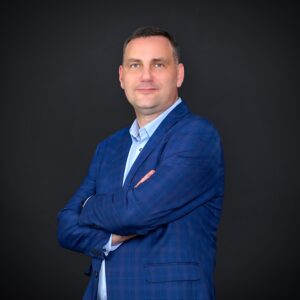 Portret mężczyzny w niebieskiej marynarce i jasnej koszuli, stojącego z założonymi rękami na ciemnym tle. Mężczyzna patrzy prosto w obiektyw z delikatnym uśmiechem.