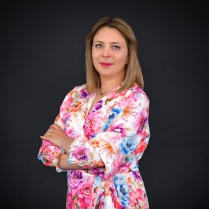 Kobieta o blond włosach, ubrana w kolorowy szlafrok w kwiaty, stoi ze skrzyżowanymi ramionami na ciemnym tle.