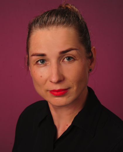 Portret kobiety o jasnej karnacji, niebieskich oczach i czerwonych ustach, ubranej w czarną koszulę, na tle fioletowej ściany.