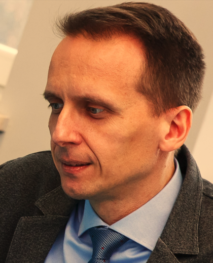 Portret mężczyzny w średnim wieku, ubranego w szarą marynarkę i jasnoniebieską koszulę z krawatem. Mężczyzna patrzy w dół i w lewo, ma krótkie, ciemne włosy.