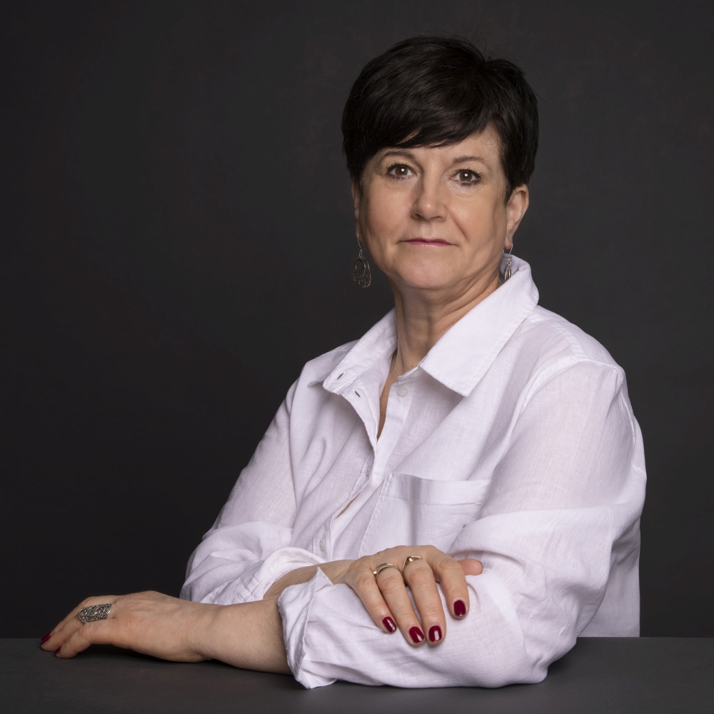 Portret kobiety w średnim wieku z krótkimi, ciemnymi włosami, ubranej w białą koszulę, siedzącej przy ciemnym stole z rękami skrzyżowanymi. Kobieta ma na sobie biżuterię i patrzy prosto w obiektyw.