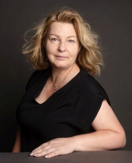 Portret uśmiechniętej kobiety w średnim wieku z blond włosami, ubranej w czarną bluzkę, opierającej się o ciemny blat. Kobieta patrzy prosto w obiektyw, a jej włosy są lekko rozwiane.