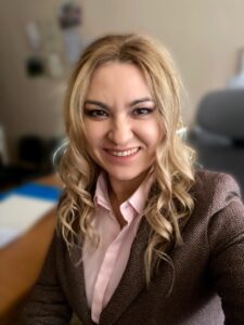 Portret uśmiechniętej kobiety o blond włosach, Ewy Sieradzkiej, siedzącej w biurze. Ma na sobie różową koszulę i brązową marynarkę, patrząc prosto w obiektyw.
