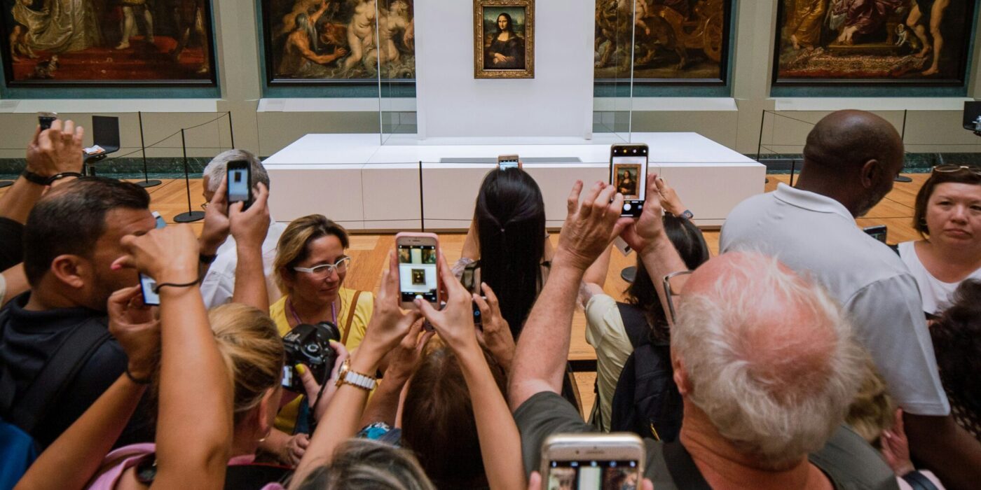 Tłum ludzi w muzeum Louvre robi zdjęcia słynnego obrazu Mona Lisa, trzymając smartfony w górze. W tle widać inne duże dzieła sztuki na ścianach galerii.