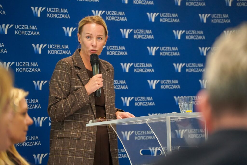dr Joanna Lizut prorektor Uczelni Korczaka w brązowej marynarce przemawia do uczestników CARESKILLS TALKS 3, trzymając mikrofon. W tle widoczne logo Uczelni Korczaka
