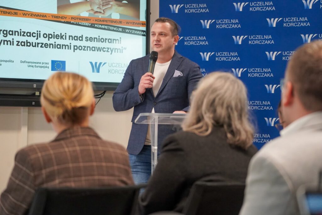Prowadzący wydarzenie Jakub Wróblewski. Mężczyzna w niebieskiej marynarce przemawia do zgromadzonych uczestników