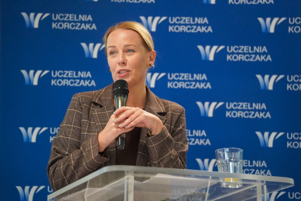 dr Joanna Lizut prorektor Uczelni Korczaka w brązowej marynarce przemawia do uczestników CARESKILLS TALKS 3, trzymając mikrofon. W tle widoczne logo Uczelni Korczaka
