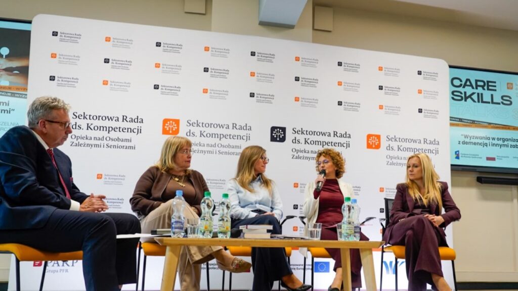 Prowadzący dyskusję i panelistki podczas rozmowy o wsparciu osób z demencją i ich opiekunów. Ujęcie ogólne na tle ścianki z logotypami Sektorowej Rady i ekranów multimedialnych.