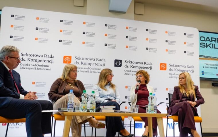 Prowadzący dyskusję i panelistki podczas rozmowy o wsparciu osób z demencją i ich opiekunów. Ujęcie ogólne na tle ścianki z logotypami Sektorowej Rady i ekranów multimedialnych.